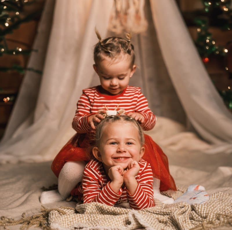 Two little girls in red-and-white striped pyjamas in front of a Christmas teepee with fairy lights