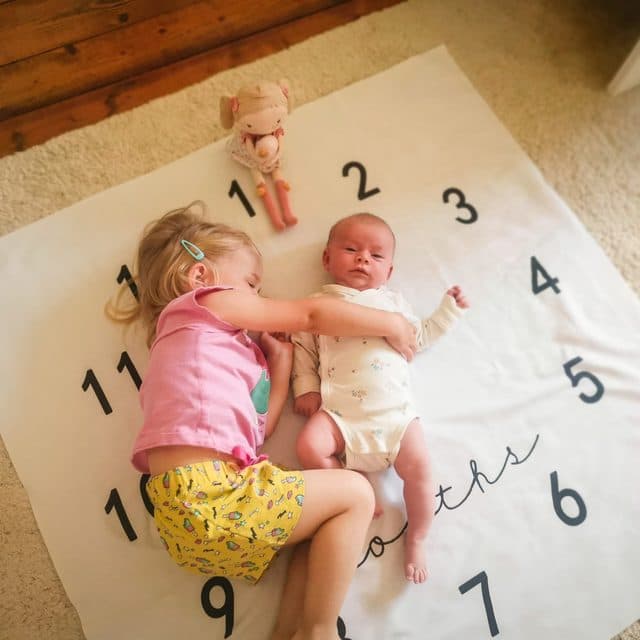 Big sister hugging a newborn on a white blanket with month numbers 1 to 12